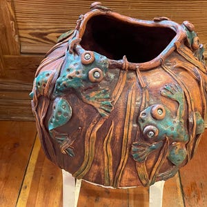 May include: A handcrafted, ceramic vase with a wide opening. The vase is a warm brown color with teal frog and aquatic plant relief details. The frogs have large, round eyes. The vase is on a white stand.