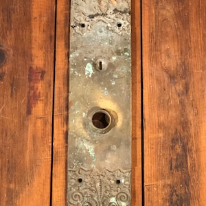 May include: Antique brass door plate with intricate floral designs and a keyhole. The plate has a worn, aged patina.
