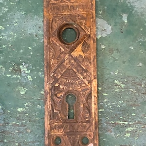May include: Antique brass door plate with intricate design and a keyhole. The plate has a worn, aged look and is rectangular in shape.