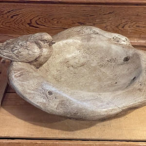 May include: A beige bird bath with a sculpted bird perched on the side. The bird bath has a shallow basin and a textured, weathered appearance, suggesting a rustic or garden theme.