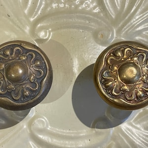 May include: Two antique brass door knobs with intricate floral designs. The knobs have a circular shape and a raised center.