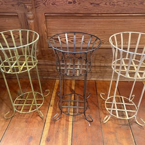 May include: Three metal plant stands with a distressed finish. The stands have a cage-like design with a round base and a flared top. One is black, and the other two are off-white.