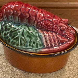 May include: A ceramic casserole dish with a lobster-shaped lid. The lid is a glossy red, with green leaf accents. The dish itself is a warm brown color with a black rim. A decorative kitchen item.
