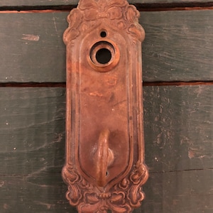 May include: Antique brass door plate with ornate floral details and a keyhole. The plate has a hook on the bottom.