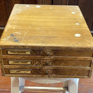 May include: A wooden drawer unit with four drawers. The drawers have brass pulls and are labeled with handwritten text. The unit is sitting on a white stool.