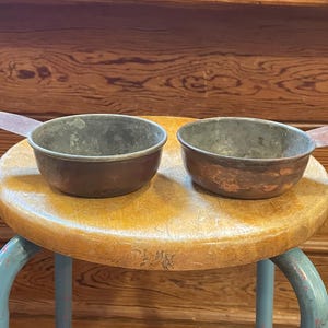 May include: Two antique metal measuring cups with handles. The cups are a tarnished copper color, and sit on a worn wooden stool with blue legs. The handles are flat and extend from the side of each cup.