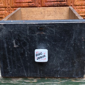 May include: Black wooden drawer with a white porcelain knob with red arrows and the text "BAC. JUNIP."