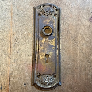 May include: Antique brass door plate with a keyhole and a circular knob opening. The rectangular plate features floral designs at the top and bottom. The brass has a weathered, aged appearance with areas of discoloration.