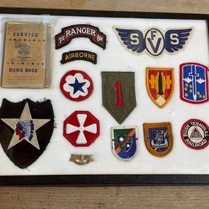 May include: A framed collection of military patches and a memo book. Patches include a black shield with a white star and Native American head, a red and white Ranger patch, and a patch with the text "AIRBORNE".