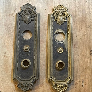 May include: Two antique brass elevator buttons with ornate frames and a shield design. Each button has three round openings for buttons.