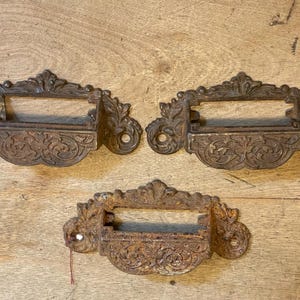 May include: Three antique bronze drawer pulls with ornate floral and scrollwork designs. Each pull has a rectangular opening for a label and decorative flourishes on the sides. The pulls are aged with a rustic, weathered appearance.