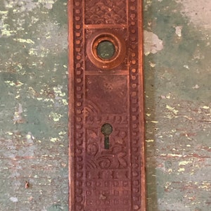 May include: Antique brass door plate with intricate floral and geometric designs. The plate has a keyhole and a round hole for a doorknob.