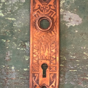 May include: Antique brass door plate with intricate floral and geometric designs. The plate has a keyhole and a round knob hole.