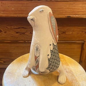 May include: A cream-colored ceramic rabbit figurine with decorative patterns in black, red, and brown. The rabbit has long ears, a detailed face, and is posed upright. The figurine is placed on a light-colored wooden stool.