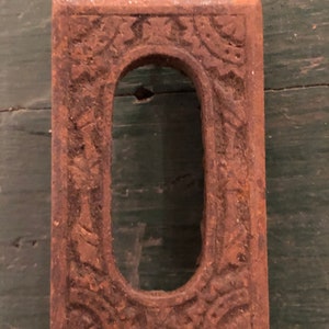 May include: A rusty metal keyhole cover with an ornate design. The cover is rectangular with a keyhole in the center. The design is intricate and detailed, with a floral motif.