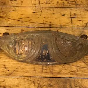 May include: A vintage, ornate, metal drawer pull with a decorative, scroll-like design. The pull is a dark, aged green color with rust spots and has a slightly curved shape.