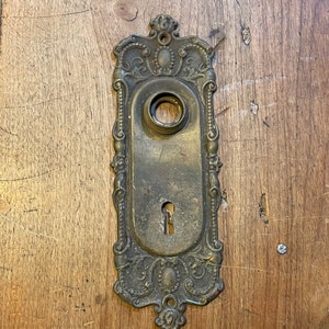 May include: Ornate brass door plate with a keyhole and a round hole for a knob. The plate has a decorative design with intricate details.
