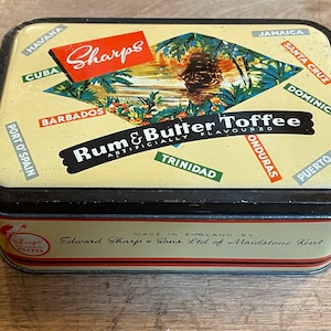 May include: A vintage tin box for Sharps Rum & Butter Toffee. The box is yellow with a red and white label. The label features a ship sailing through a tropical scene with palm trees and the text "Sharps Rum & Butter Toffee" in black letters. The box is decorated with the names of various Caribbean islands in a colorful font.