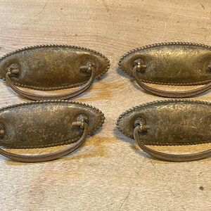 May include: Four antique brass drawer pulls with oval backplates and curved handles. The pulls have a beaded edge and a tarnished finish, suggesting age and use. These hardware pieces are likely for a dresser or cabinet.