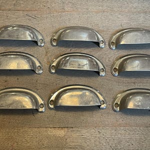May include: Nine vintage, silver-toned metal drawer pulls arranged on a wooden surface. Each pull has a half-moon shape with two screw holes. The pulls have a slightly aged appearance.
