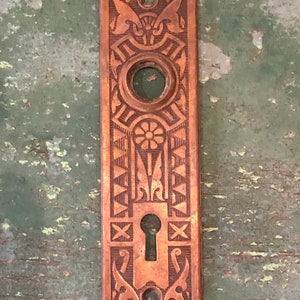 May include: Antique brass door plate with an ornate design featuring a keyhole and a round knob. The plate has a dark brown patina and is in good condition.