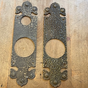 May include: Two antique brass door plates with a hammered finish and ornate designs. Each plate has a circular cutout for a door knob.