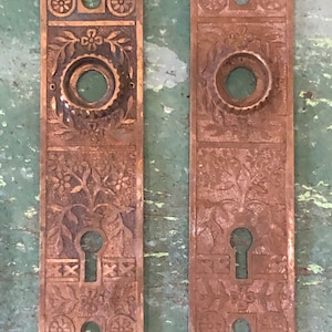 May include: Two antique brass door plates with intricate floral designs and keyholes. The plates have a worn, aged patina.