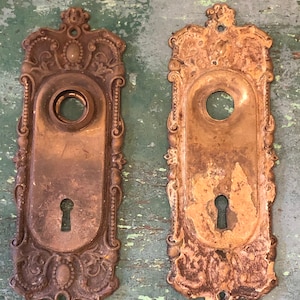 May include: Two antique brass door plates with ornate designs. The plates have a keyhole and a round opening for a knob. The plates are worn and have a patina finish.