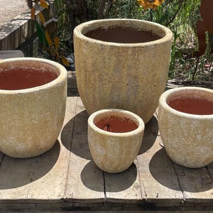 May include: Four beige-colored, textured ceramic planters of varying sizes. The planters have a rounded shape with a wide opening and a reddish-brown interior. They are arranged on a weathered wooden surface, suggesting an outdoor setting.