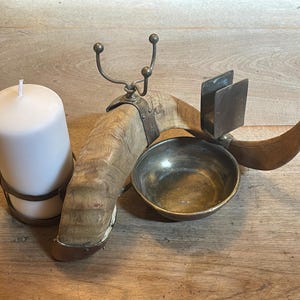 May include: An antique candle holder with a white pillar candle, a wooden base, and metal accents. The holder features a small bowl, a curved horn-like element, and decorative metal hooks. The candle is held in a metal ring.