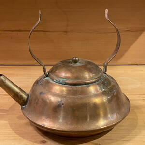 May include: A vintage copper teapot with a hinged lid and a curved handle. The teapot has a worn, aged patina and a spout on the side.