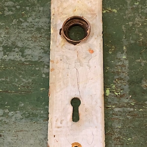 May include: White metal door plate with a keyhole and two round holes. The plate has a distressed, aged look with cracks in the paint.