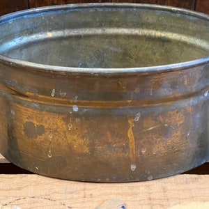 May include: A large, round, brass planter with a worn, aged patina. The planter has a slightly oval shape and a smooth, shiny surface.