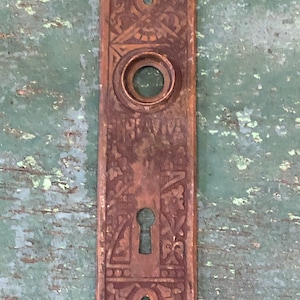May include: Antique brass door plate with intricate floral and geometric designs. The plate has a keyhole and a round hole for a door knob.