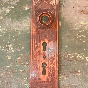 May include: Antique brass door plate with intricate engraved designs, including a keyhole and a circular knob. The plate has a worn, aged appearance.