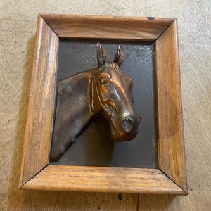 May include: A framed metal relief sculpture of a horse's head. The horse is depicted in profile, with detailed features and a copper-colored finish. The frame is made of wood with a natural finish, and the background is a dark gray color.