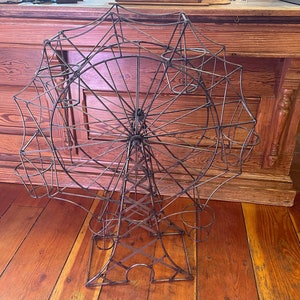 May include: A decorative metal Ferris wheel planter with multiple circular openings for plants. The structure is made of thin, dark metal rods, and stands on a base with decorative cutouts. The background features a wooden surface.