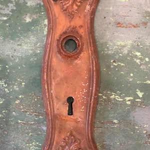 May include: A vintage, rusty, copper-colored door plate with a keyhole and decorative floral accents.