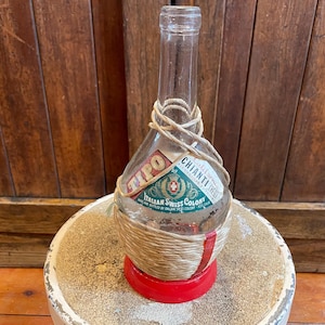 May include: A clear glass bottle with a straw-wrapped neck and a red base. The bottle has a label that reads "Tipo Chianti Italian Swiss Colony".