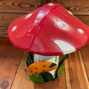 May include: A red and white mushroom-shaped metal garden sculpture with green leaves and a yellow and orange flower.