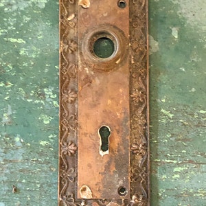May include: Antique brass door plate with ornate floral design and a keyhole. The plate has a worn, aged patina.