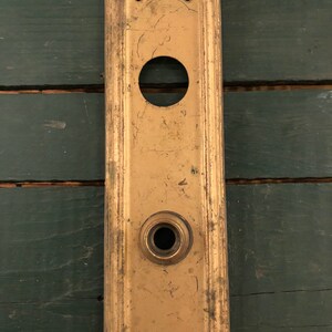 May include: A vintage brass door plate with a keyhole and a round hole for a door knob. The plate has a worn, aged look and is a rectangular shape.