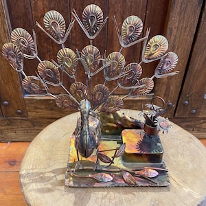 May include: A copper and brass peacock music box with a detailed design. The peacock has a spread tail of feathers and is standing on a base with a music box mechanism. The music box is decorated with flowers and leaves.
