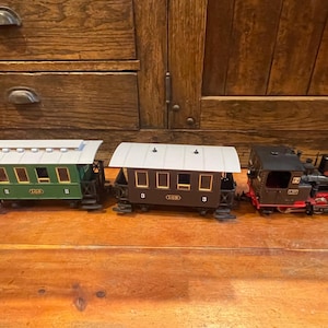May include: A model train set featuring a black locomotive, a green passenger car, and a brown passenger car. The train cars have white roofs and are set on a wooden surface.