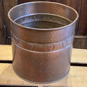 May include: A hammered copper planter with a decorative heart design around the top rim. The planter is a cylindrical shape and has a rustic, aged look.