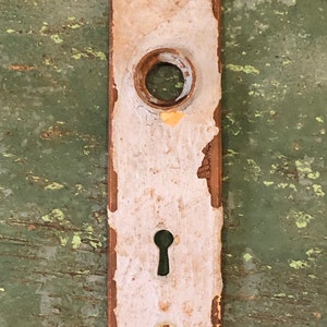 May include: A vintage white and brown door knob backplate with a keyhole and a round knob hole. The backplate is slightly chipped and worn.