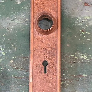 May include: A vintage brass door plate with a decorative swirl pattern. The plate has a keyhole and a round opening for a door knob.