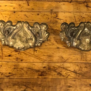May include: Two ornate, gold-toned metal drawer pulls with a floral design. The pulls are on a wooden surface.