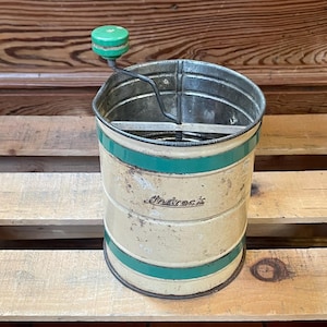 May include: Vintage green and white metal flour sifter with a crank handle. The sifter has a green stripe around the top and bottom and the word "Antrock" printed on the side.