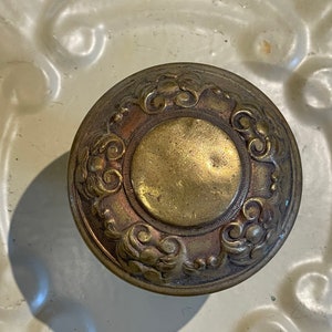 May include: A round, brass door knob with a decorative design of swirling patterns and a central circle. The knob has a worn, antique finish.